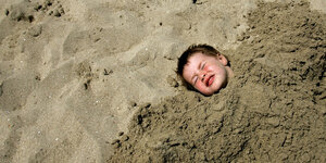 Ein Junge hat sich in Sand eingebuddelt, nur noch der Kopf ragt raus.