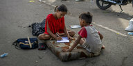 Ein Mädchen schreibt in ein Heft, sie sitzt mit einem Jungen auf einer Matratze auf der Straße