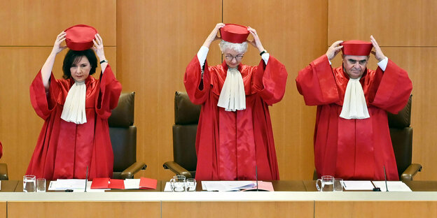 drei Menschen in roten Roben vor holzvertäfelten Wänden greifen an ihre rote Kopfbedeckung