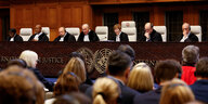 Sieben Richter:innen sitzen in ihren Roben an einem langen Tisch aus dunklem Holz auf dem das Logo der Vereinten Nationen prangt