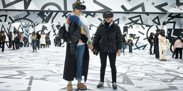 Zwei Frauen halten sich an den Händen und tragen VR Brillen, im Hintergrund viele Menschen mit VR Brillen