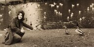 Eine junge dunkelhaarige Frau kniet an einem Weiher, um sie herum Enten