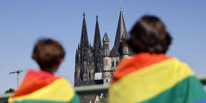 Zwei Menschen von hinten. Sie tragen Regenbogenfahnen um die Schultern. Im Hintergrund blauer Himmel und die Türme einer Kirche.