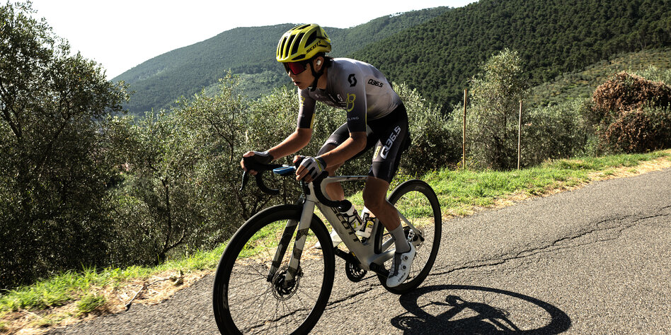 Guillermo Juan Martinez sitzt auf seinem Rennrad und strampelt einen Berg hoch, im Hintergrund bewaldete Hügel